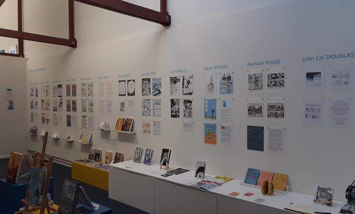 Gallery interior showing 1 long wall with artwork displayed uniformly under headings of artist names. Below on plinths and shelves are zines and books